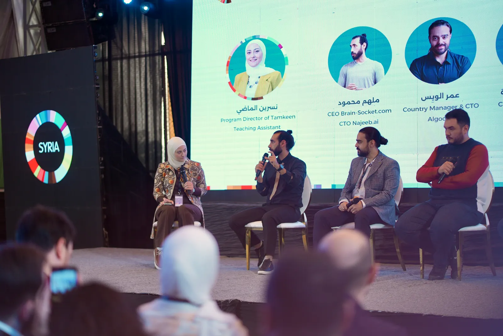The CTO Insights panel seated on stage at GEW Syria, the speaker line-up on the screen behind them